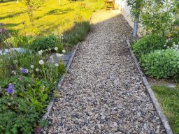 Ein Kiesweg umgeben von grünen Pflanzen und Blumen im Sonnenlicht.